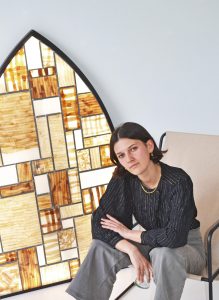 Portrait of French artist and materials researcher Loumi Le Floc'h, seated beside an arched Precious Peels panel made from dyed eggplant skin